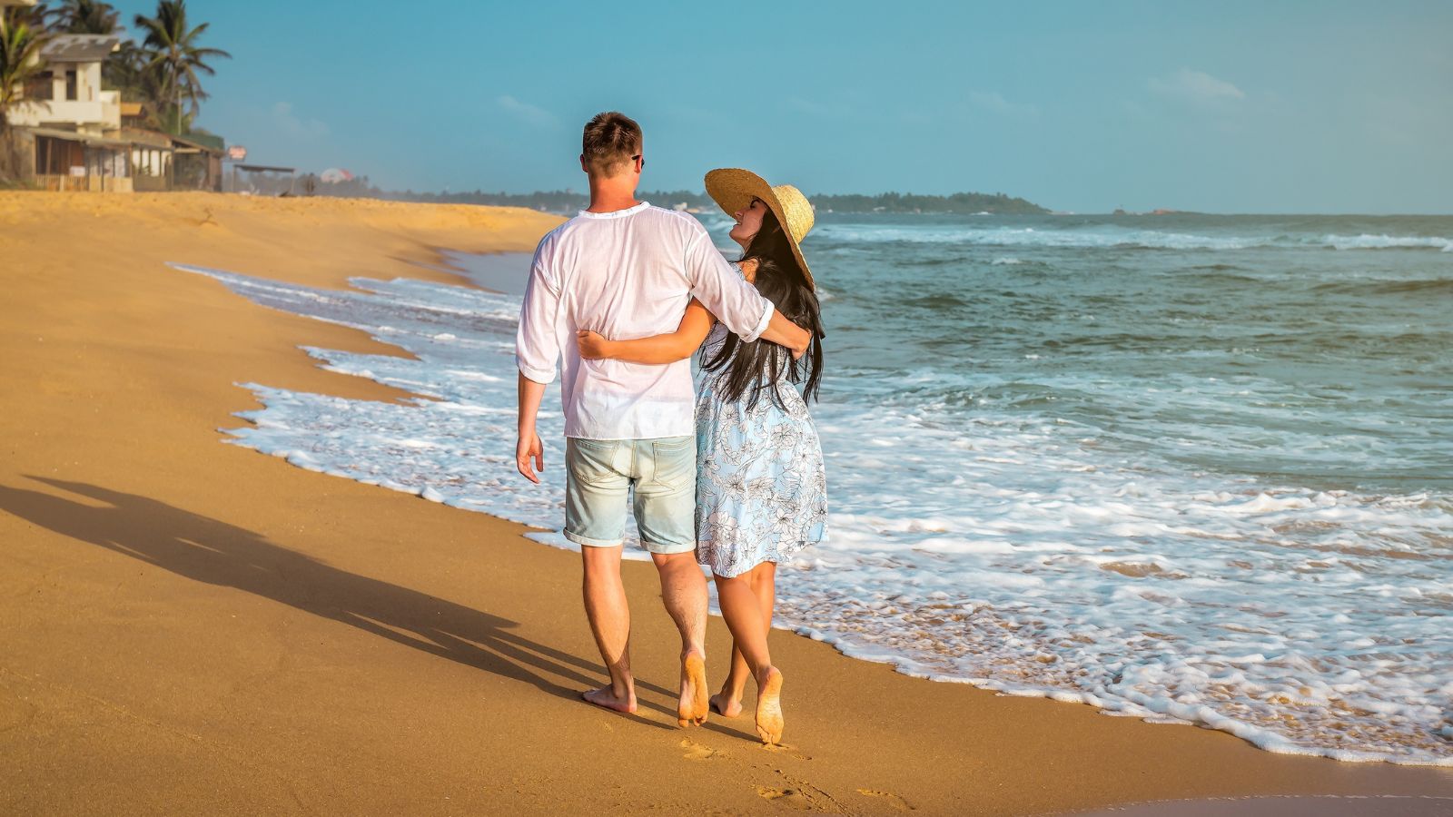 Couple walking arm in arm along a sandy beach beside the waves.